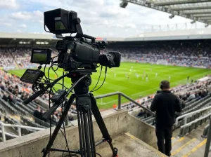 Cámara de televisión profesional grabando un partido de fútbol desde una tribuna con el campo de hierba verde en el fondo