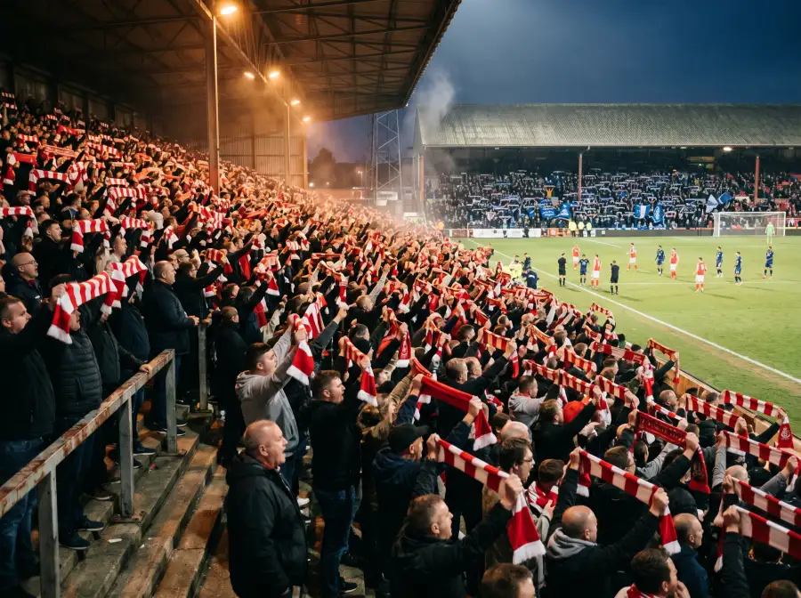 Grada de estadio inglés de fútbol abarrotada de aficionados con bufandas en alto durante un derbi de Premier League