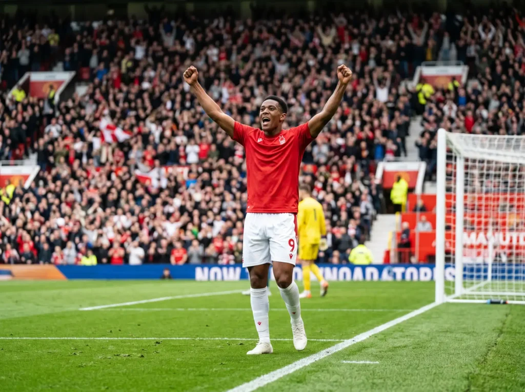 Delantero de fútbol con camiseta roja celebrando un gol dentro del área de penalti, estadio de Premier League de fondo